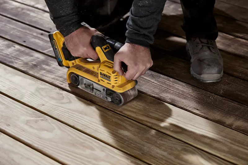 Une personne utilise une ponceuse à bande sans fil jaune DEWALT sur des planches en bois pour lisser leur surface.