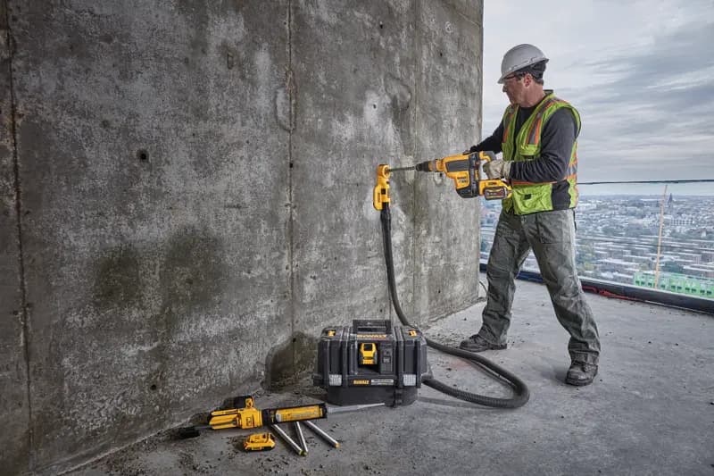 Un ouvrier du bâtiment portant un gilet de sécurité et un casque utilise une perceuse sans fil DEWALT avec un accessoire d’extraction de poussière pour percer un mur en béton. L’ouvrier se tient sur un grand bâtiment avec une vue sur la ville à l’arrière-plan. Plusieurs outils DEWALT et une mallette à outils sont posés au sol à proximité.